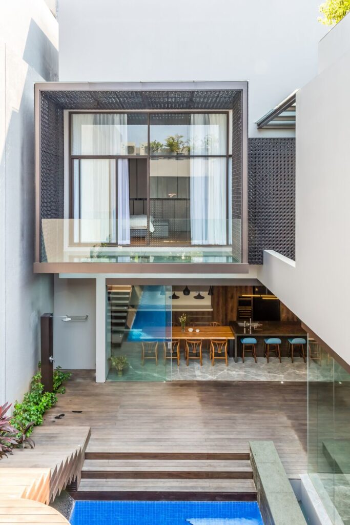 Rear view of Weave House: upper floor with a large glass window in a dark screen frame, open dining room, timber deck and pool