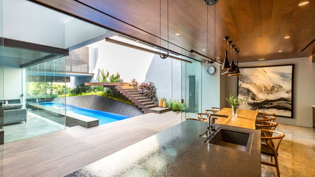 Weave House dining room opening onto the pool and garden through glass walls, with a dark kitchen counter