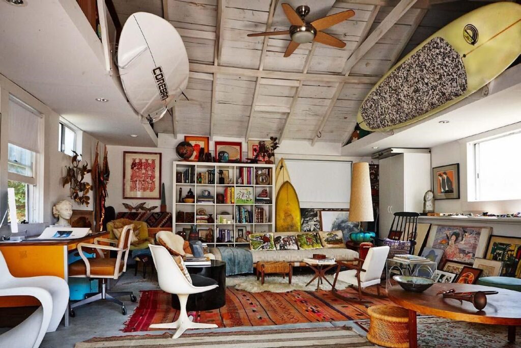 Eclectic living room mixing vintage and modern furniture, with surfboards on the ceiling, bookshelves and colourful rugs