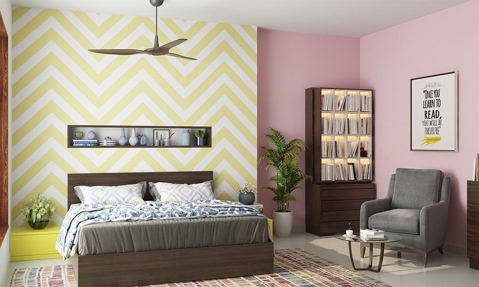 Pastel bedroom with a yellow chevron feature wall, pink walls, a grey armchair and a bookcase