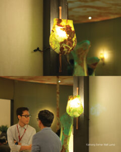 Two views of the Kantung Semar wall lamp in Golden Green onyx, lit on a dark pillar beside two visitors talking
