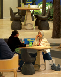 Two views of the Kantung Semar dining table with a Travertine Onyx base and teak top, one with visitors seated at it