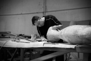 Eugenio Hendro shaping a large carved wooden form at a workbench, in black and white