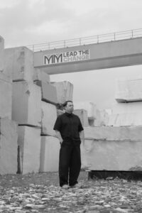 Designer Eugenio Hendro standing among stacked marble blocks beneath an MM Galleri 'Lead the Change' sign, in black and white